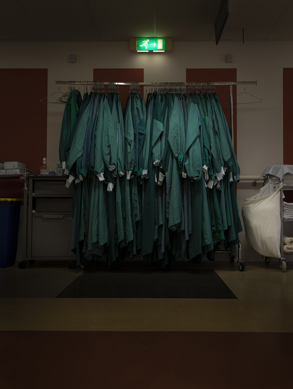 Foto van Tahne Kleijn: Een kledingrek vol groene operatiejassen staat in een ziekenhuisruimte, onder een verlicht nooduitgangsbord
