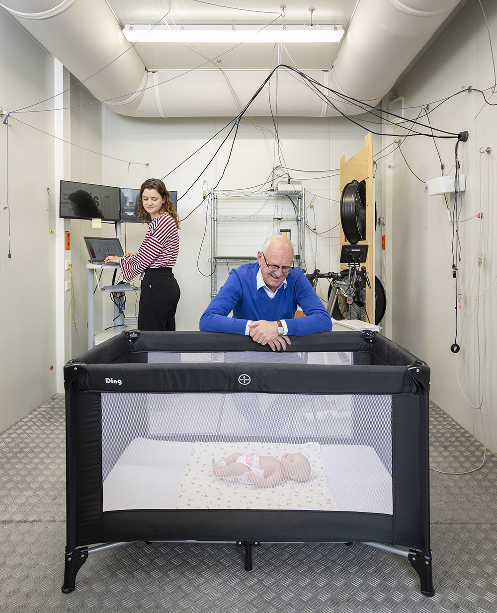 Onderzoeksruimte met meetapparatuur waarin een baby in een zwart reisbedje ligt. Op de voorgrond leunt een oudere man over het bedje, terwijl op de achtergrond een vrouw achter een laptop werkt. Kabels en sensoren hangen door de ruimte.